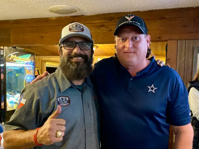 Two men in caps pose together, one giving a thumbs-up.