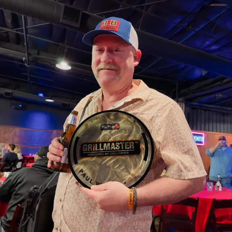 A man in a cap holds a round Grillmaster award indoors.