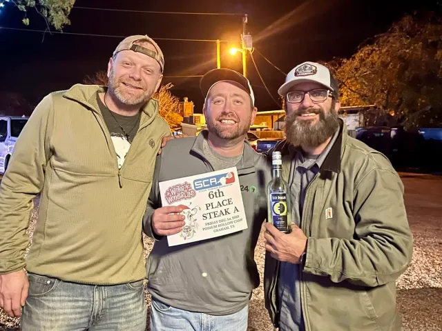 Three men pose outdoors at night with a cookoff certificate and trophy.