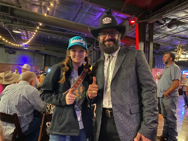 A woman and a man in a black cowboy hat give thumbs-up gestures indoors.