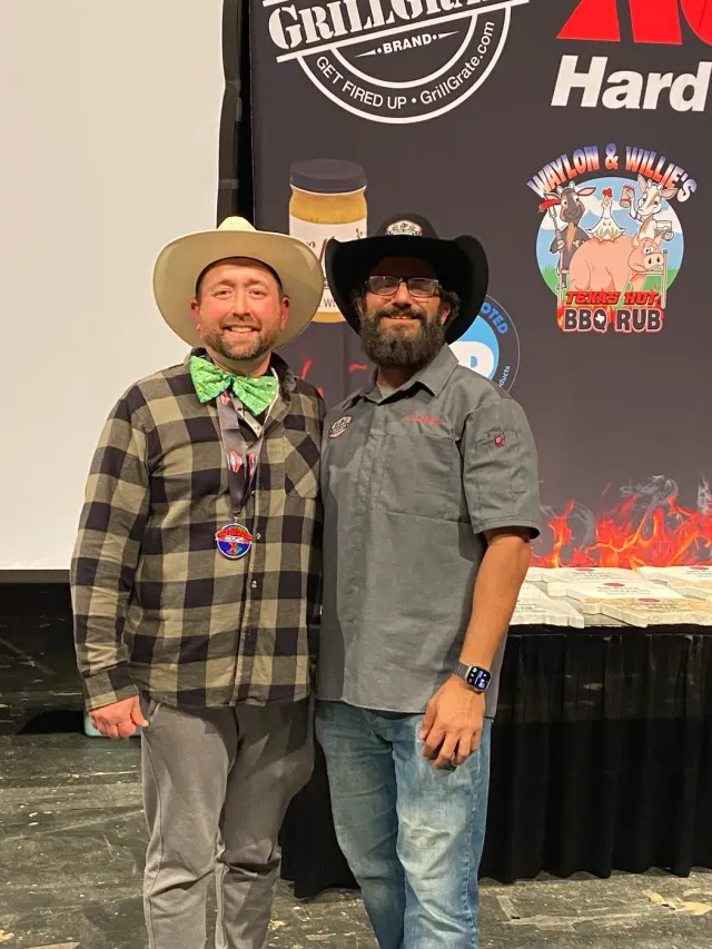Two men in cowboy hats pose in front of a cookoff event banner.
