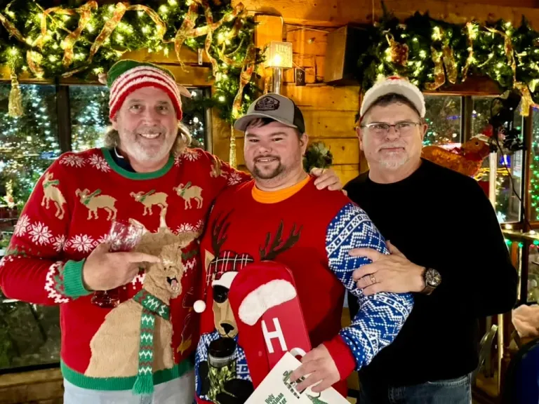 Three men in festive clothing pose indoors beneath holiday decorations.