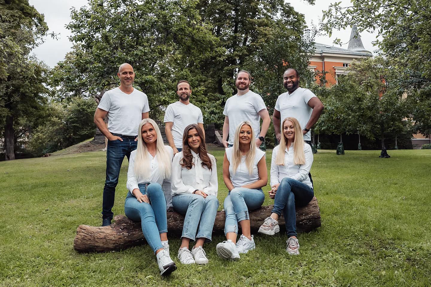 Åtte voksne personer, fire kvinner sitter på en trestamme i gressplen, fire menn står bak dem under trær.