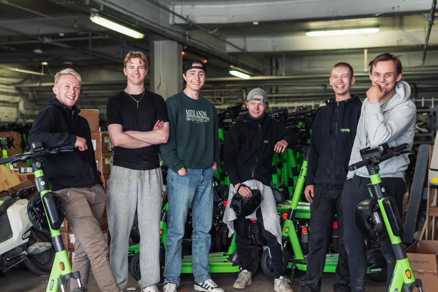 Seks unge menn smiler og står med grønne elektriske sparkesykler i et lager.
