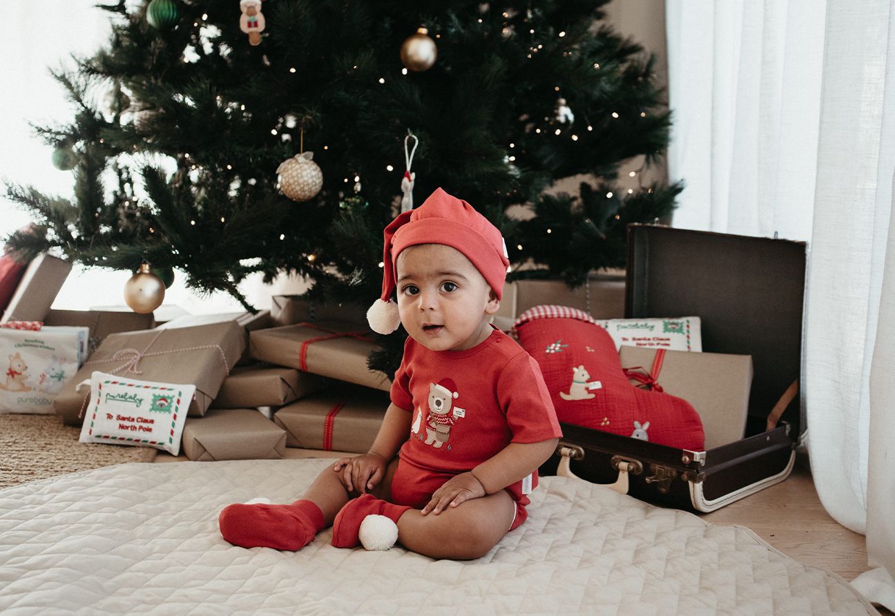 baby sitting in front of a Christmas tree wearing a Purebaby Christmas bodysuit