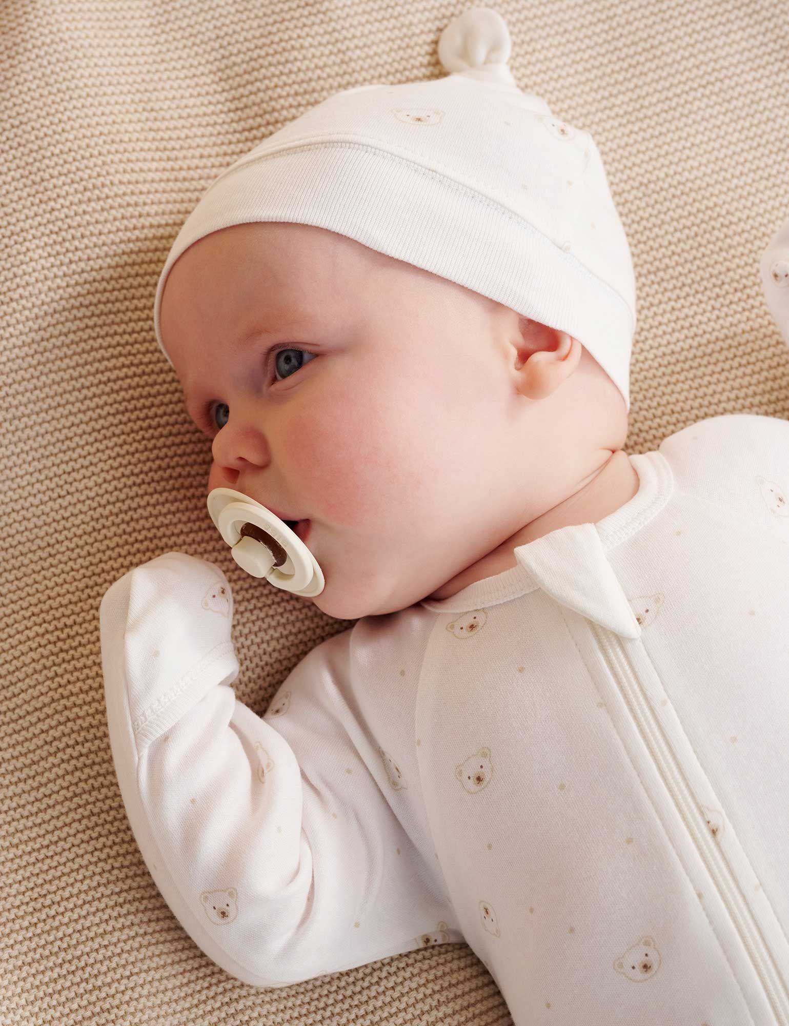 Baby wearing white onesie and hat with a white dummy in mouth