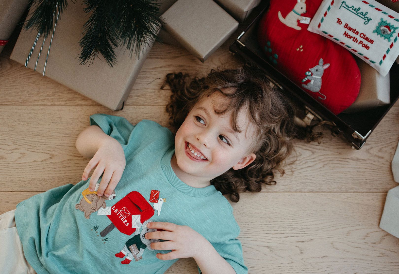Toddler wearing green Purebaby Christmas tee laying next to Christmas presents