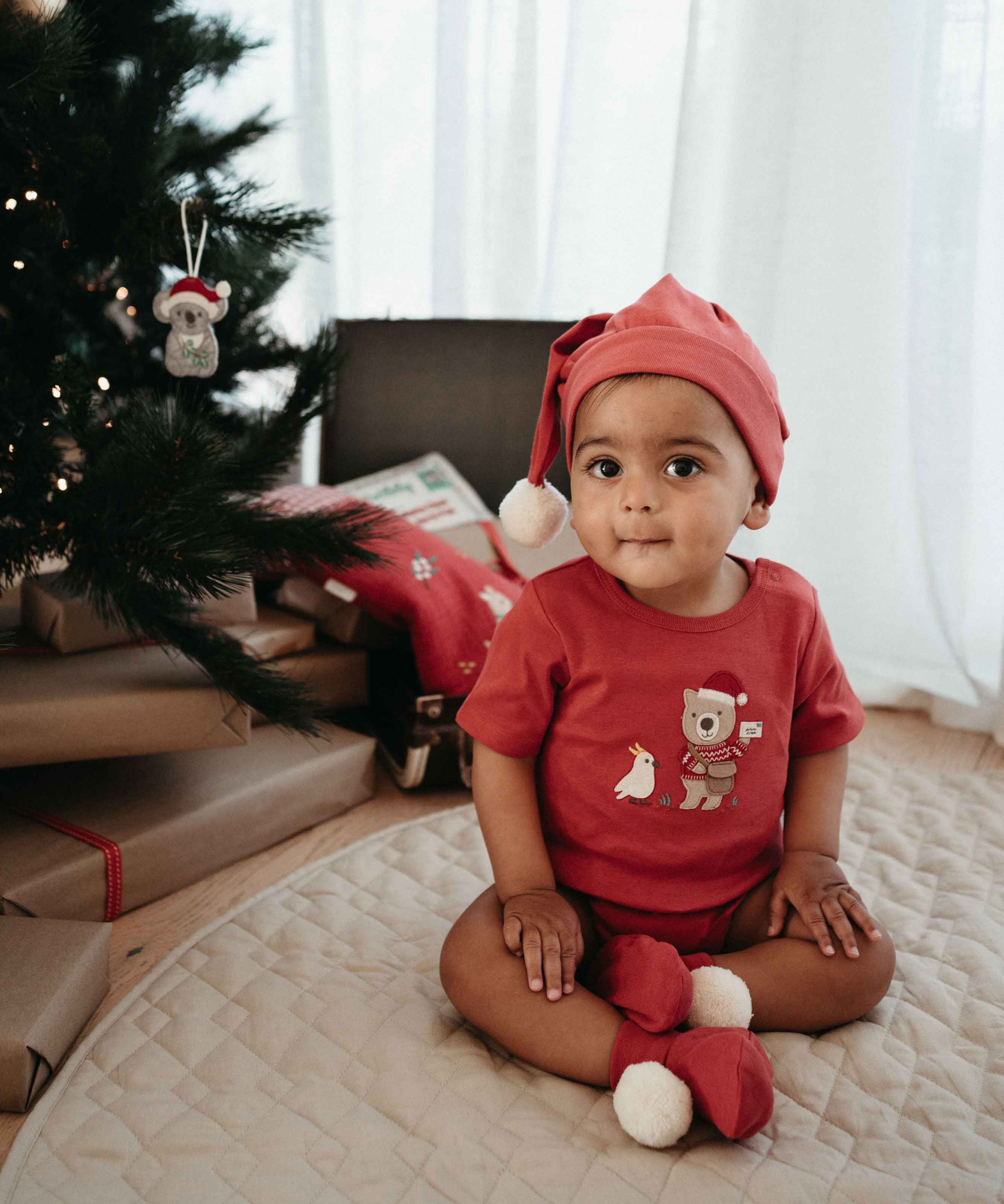 Baby wearing Postie Santa Bodysuit and hat sitting by the tree