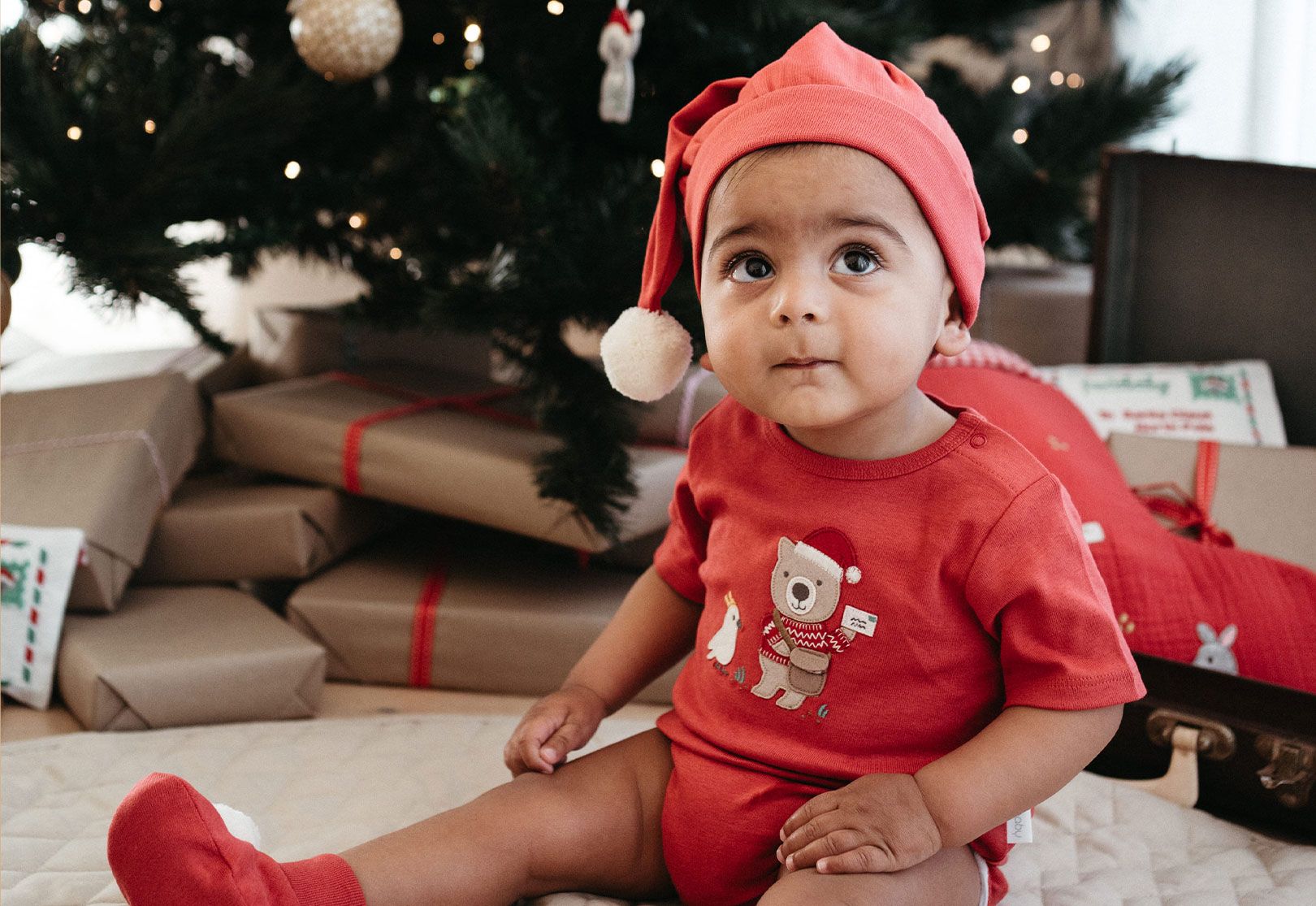 Baby sitting next to Christmas tree wearing red Purebaby bodysuit