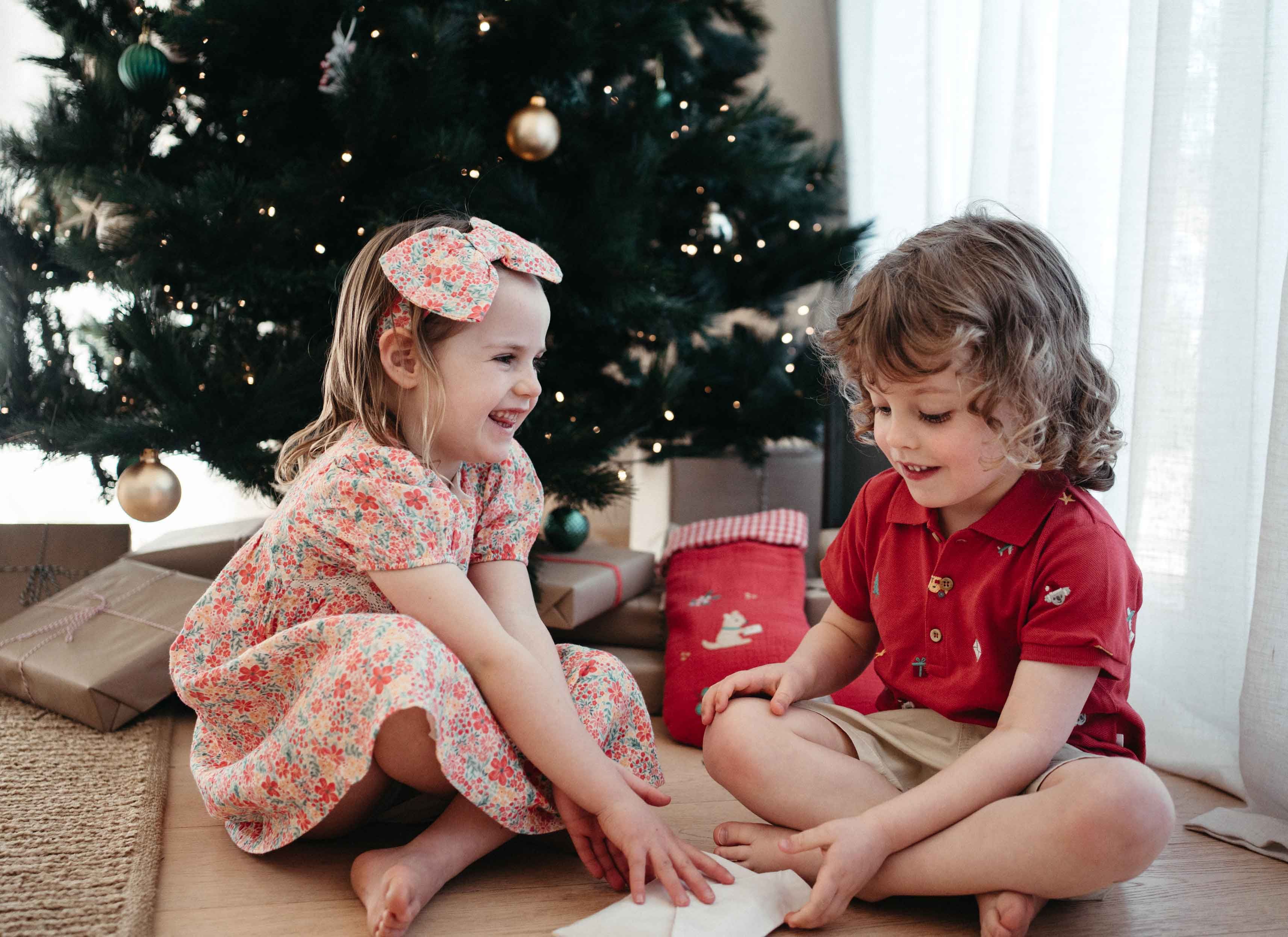 Siblings in Purebaby Christmas outfits sitting in front of Christmas tree