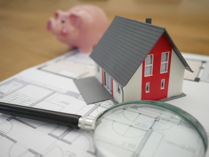 Miniature house model with a piggy bank and magnifying glass on blueprints, warm wood tones — lendsimpl guide to income needed for a mortgage in Toronto