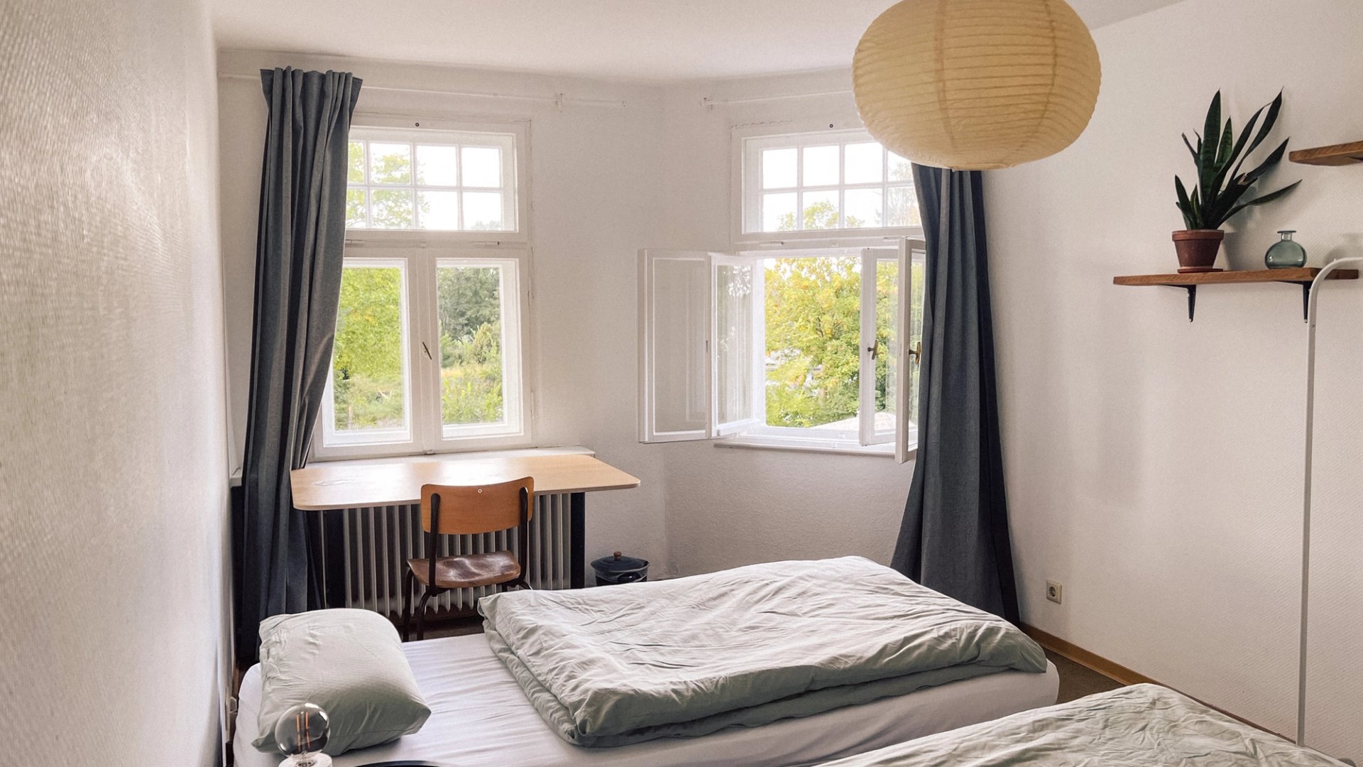 Zweibettzimmer mit Papierlampe, Kleiderstange, Schreibtisch am Fenster und Pflanzen in der Gruppenunterkunft Neupitz