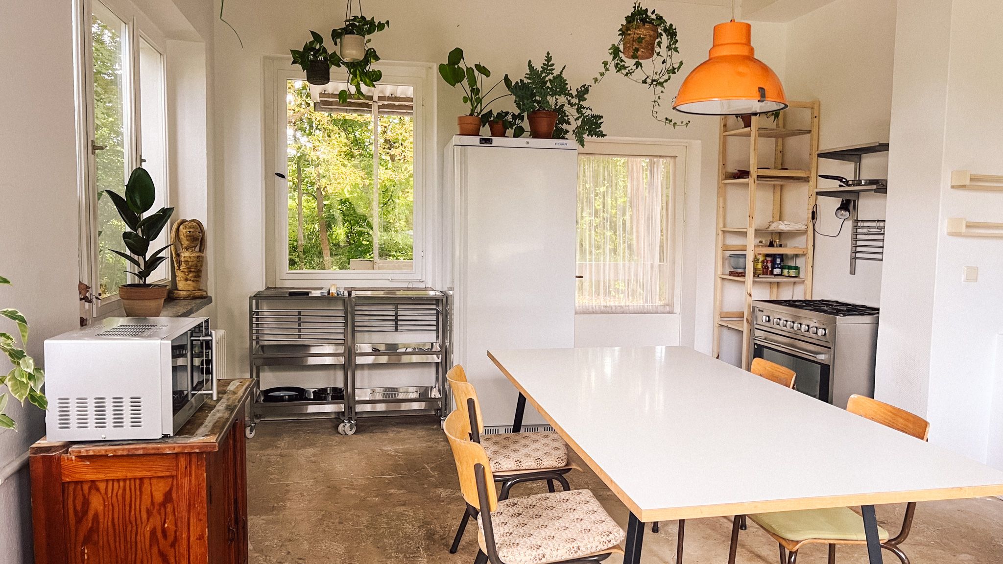 Neue Gemeinschaftsküche im Wintergarten mit langem weißen Tisch und orangefarbener Hängelampe