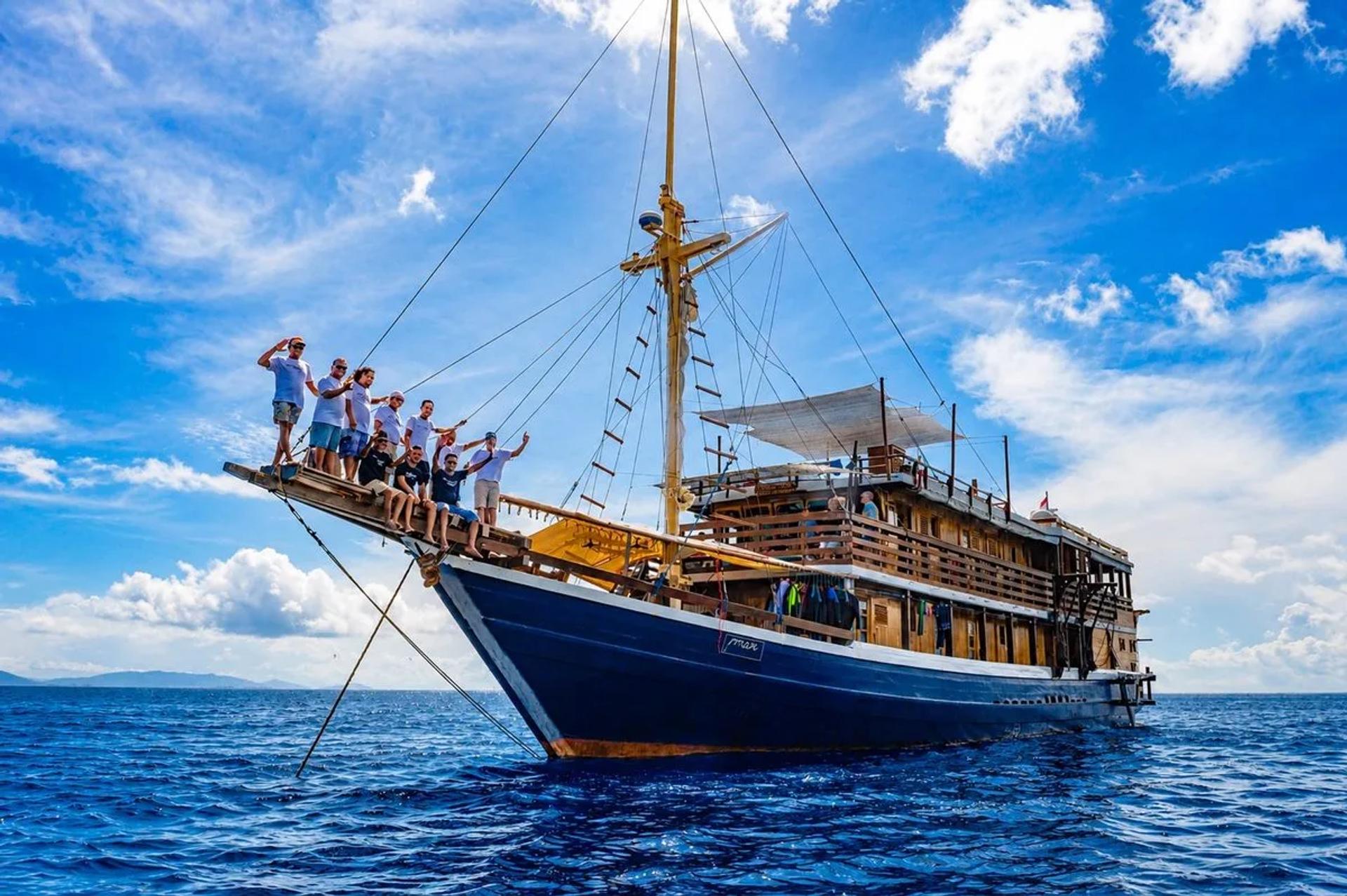 Mari crew standing on the bowsprit of the phinisi under a clear blue sky