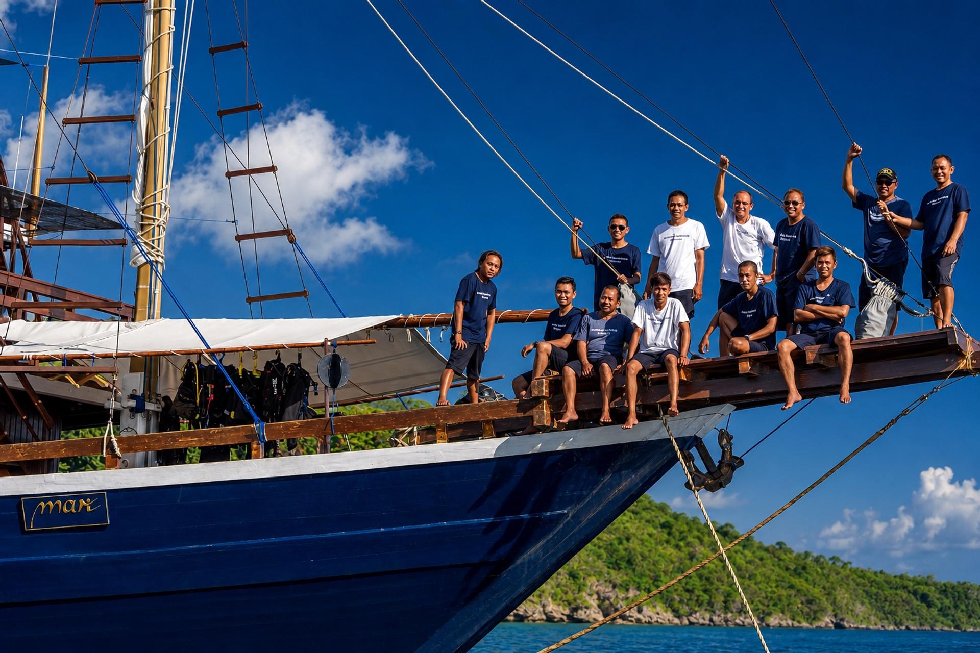 The crew lined along the bowsprit of a blue-hulled phinisi, a forested island behind
