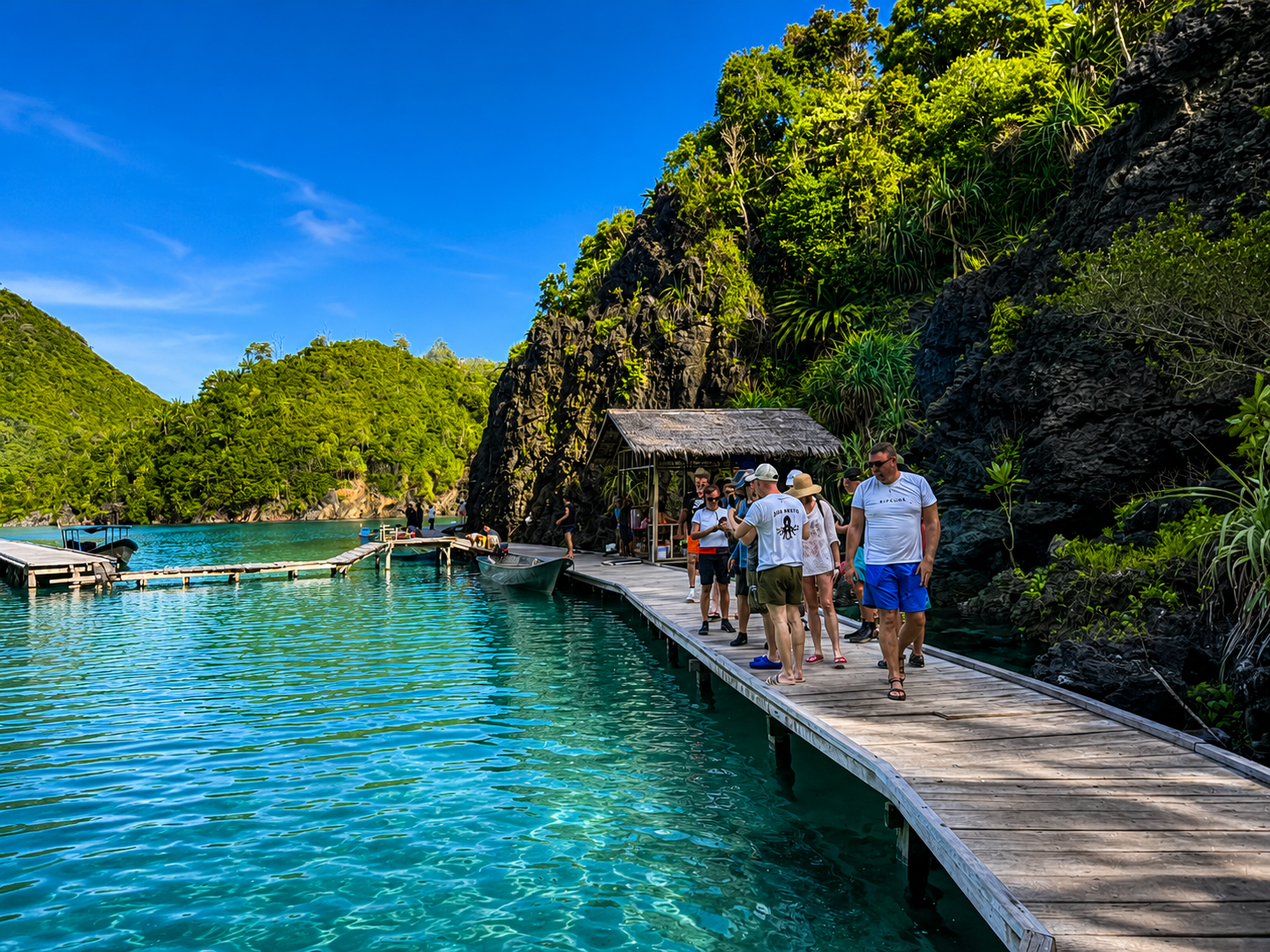 Guests on a wooden boardwalk above a turquoise bay ringed by karst