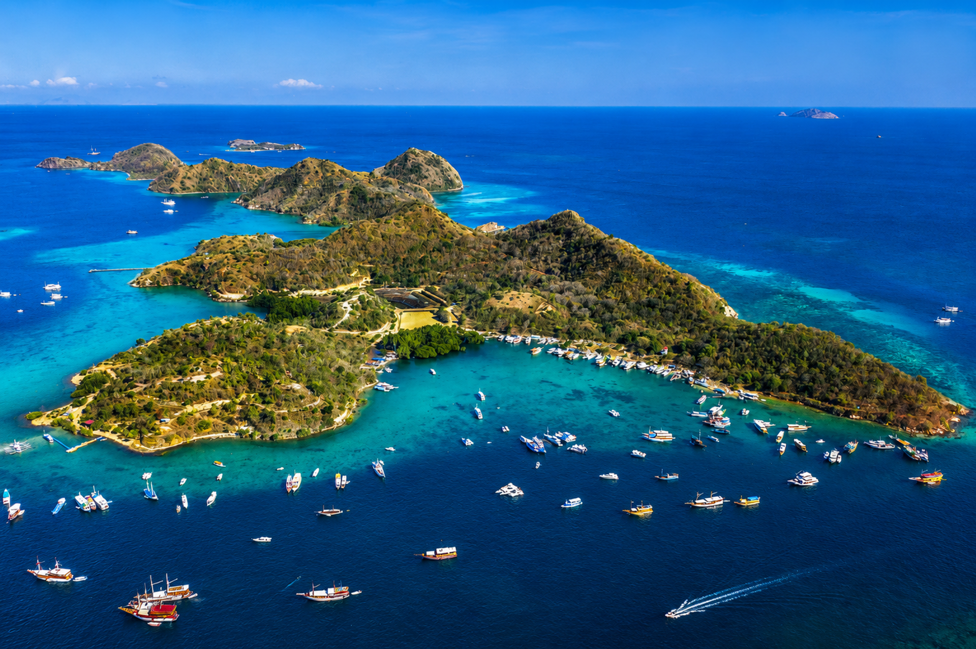 Aerial view of a green island ringed by turquoise reef with phinisi boats at anchor