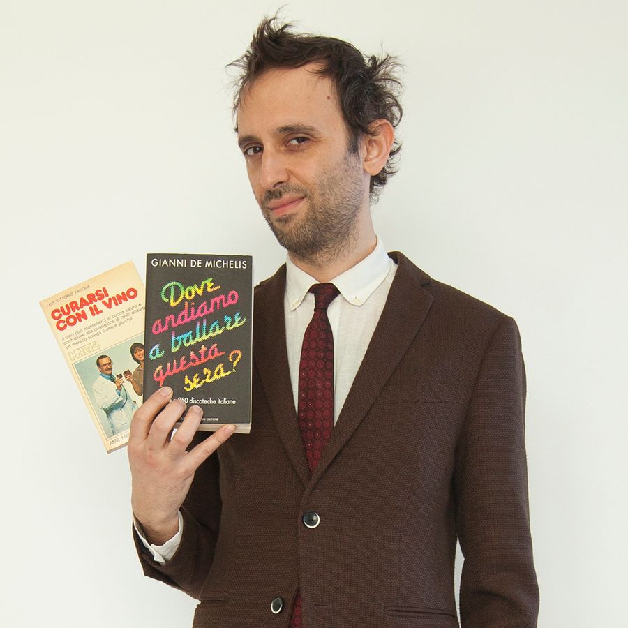 Auroro Borealo, un uomo con barba e capelli spettinati, indossa un abito marrone e una cravatta, mentre tiene in mano due libri dall'aspetto datato e dal titolo curioso.