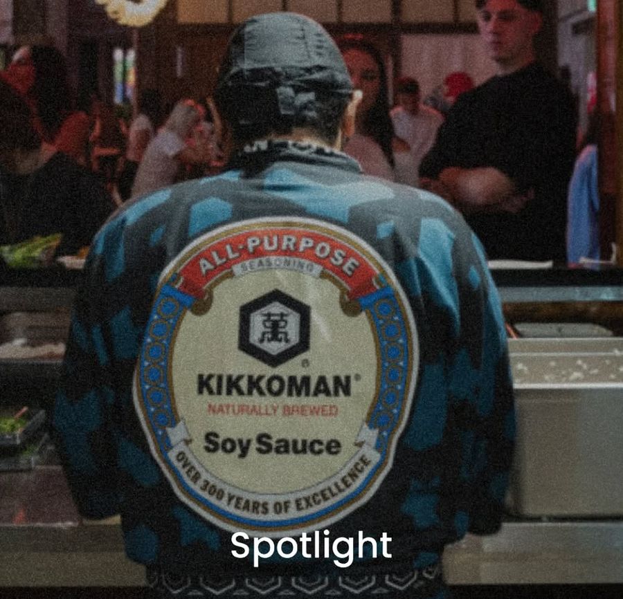Un uomo di spalle indossa un happi tradizionale giapponese con il logo Kikkoman, in un ambiente che ricrea un mercato notturno giapponese con lanterne rosse e persone sullo sfondo.