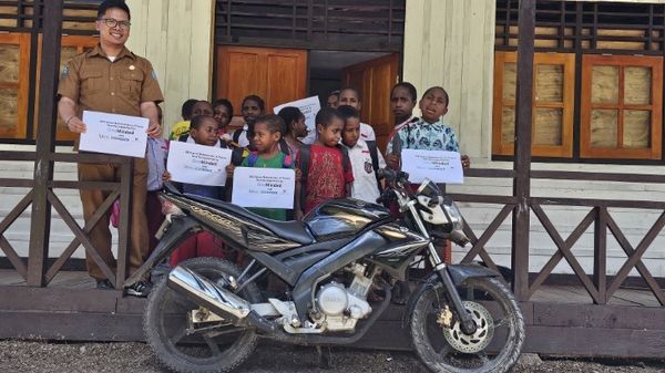 Het schoolhoofd staat naast de motor voor de school, omringd door leerlingen die kaarten omhooghouden met het project en de partners erop.