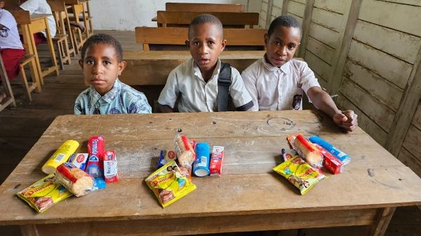 Drie jongens aan een houten schoolbank met de spullen voor hen uitgestald: tandpasta, zeep, wasmiddel en instantnoedels.