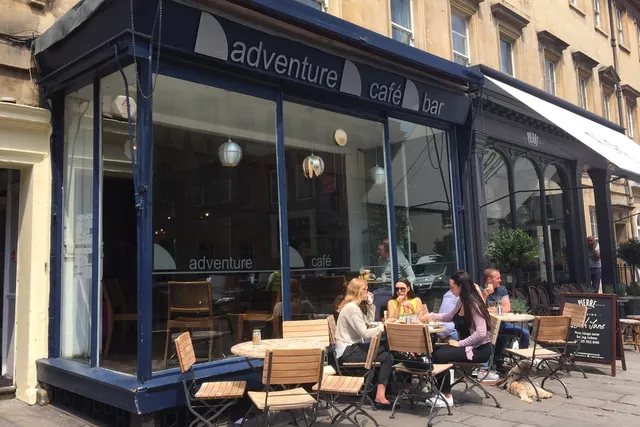 Adventure Cafe in Bath: Front view on a sunny day, featuring people seated in the outdoor seating area. The facade has large windows offering a glimpse of the vibrant interior.
