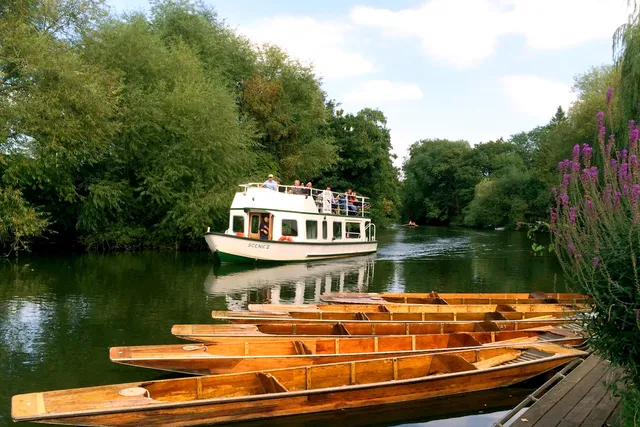 View from The Bathwick Boatman river bank in Bath, with punt boats in the foreground, and a Pulteney Cruisers tourist boat passing by, creating a picturesque riverside scene