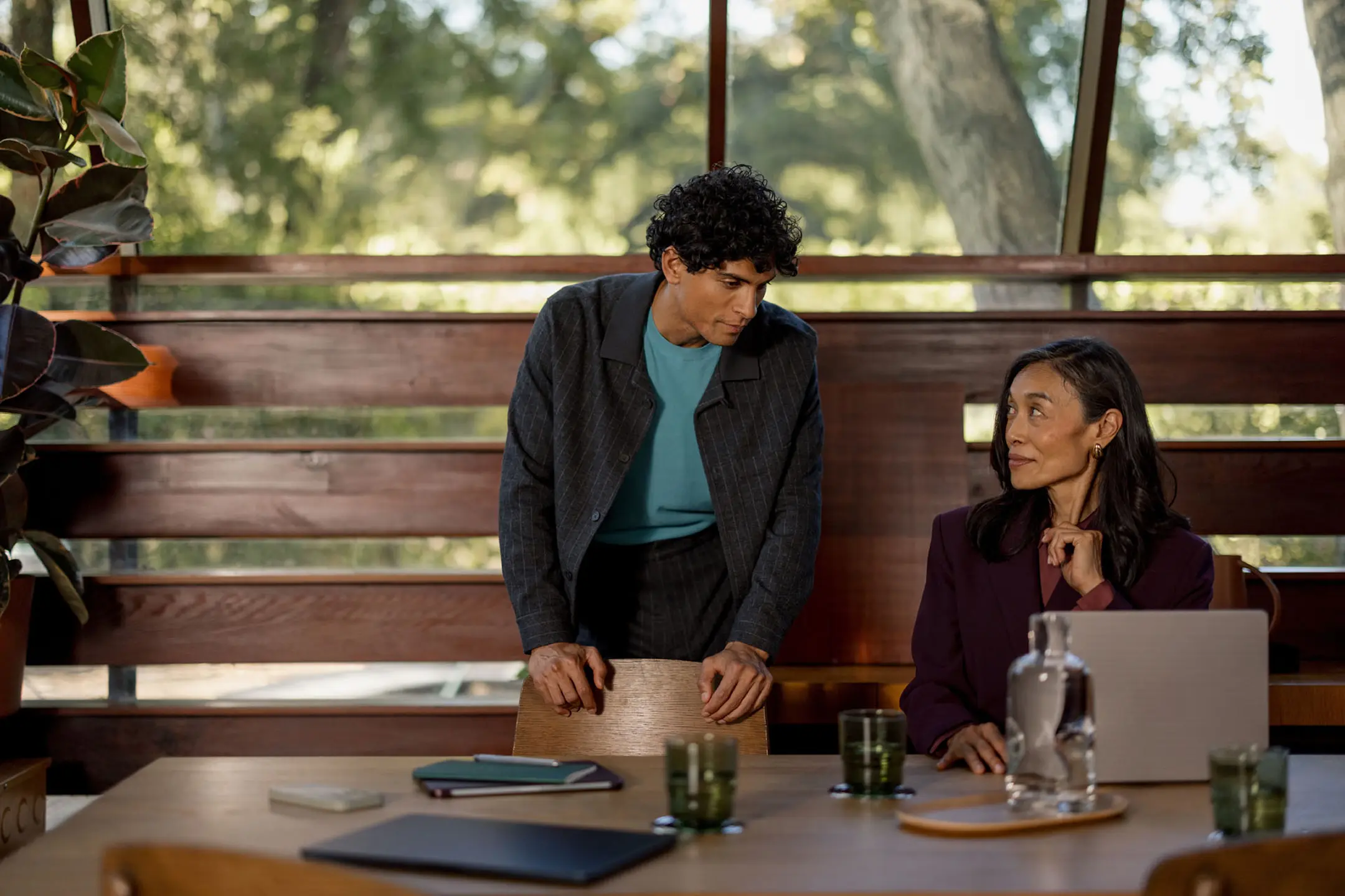 A man stands talking to a woman seated at a table in a bright modern room with large windows.