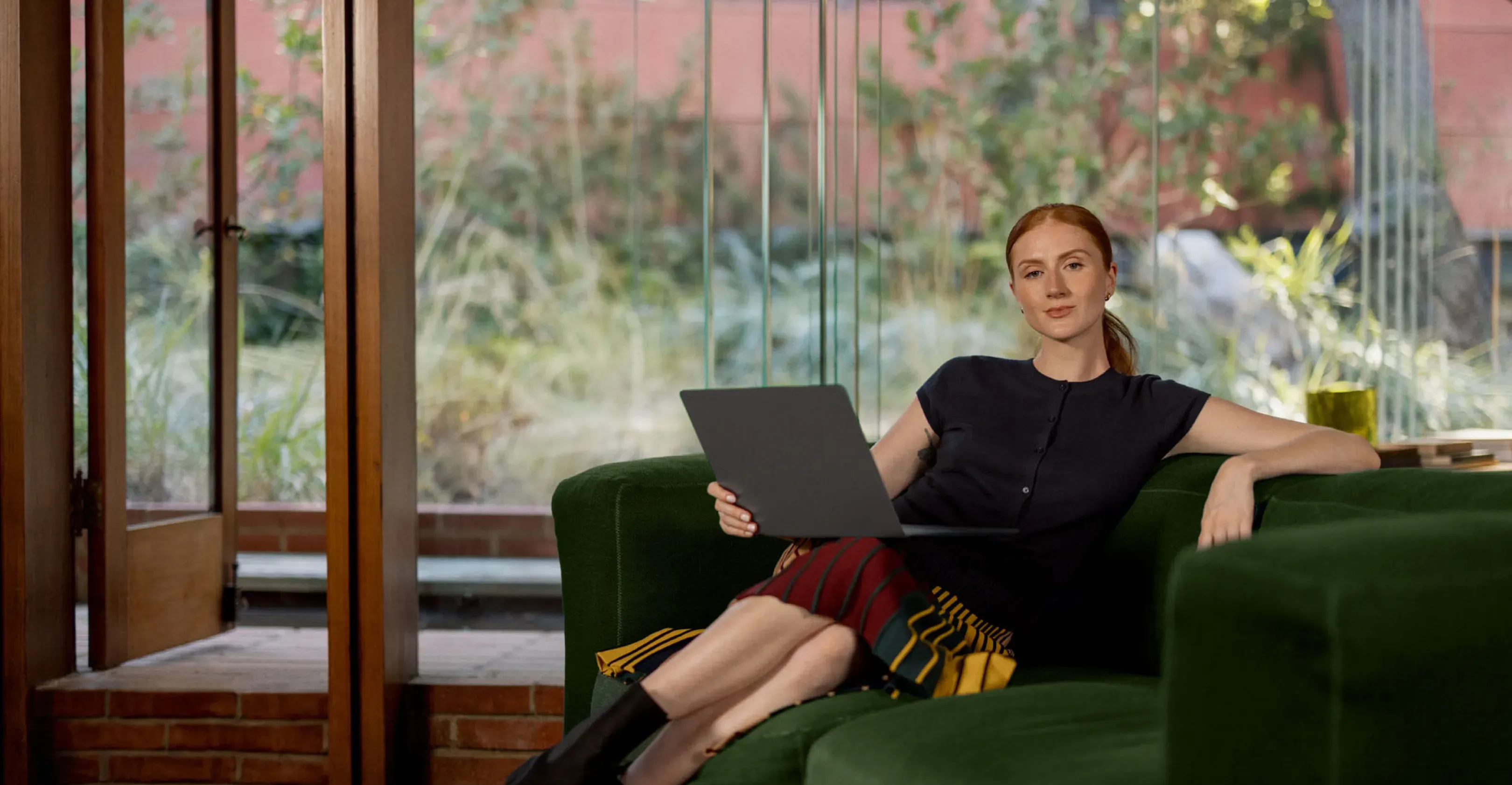 A red-haired woman sits on a green couch, holding a laptop, looking at the camera with a large window and greenery behind her.