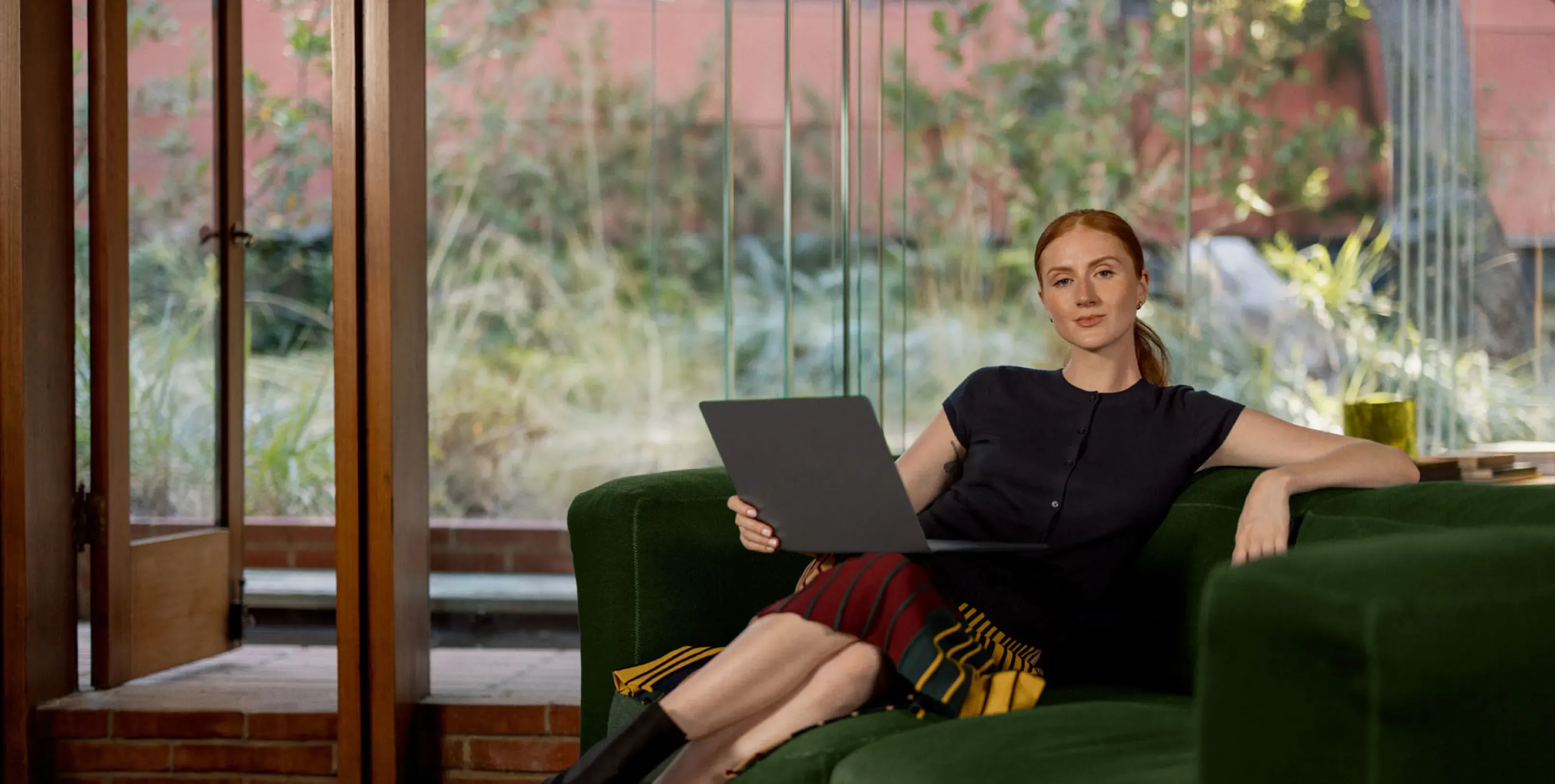 A woman with red hair sits on a green couch, holding a laptop, in a room with large windows.