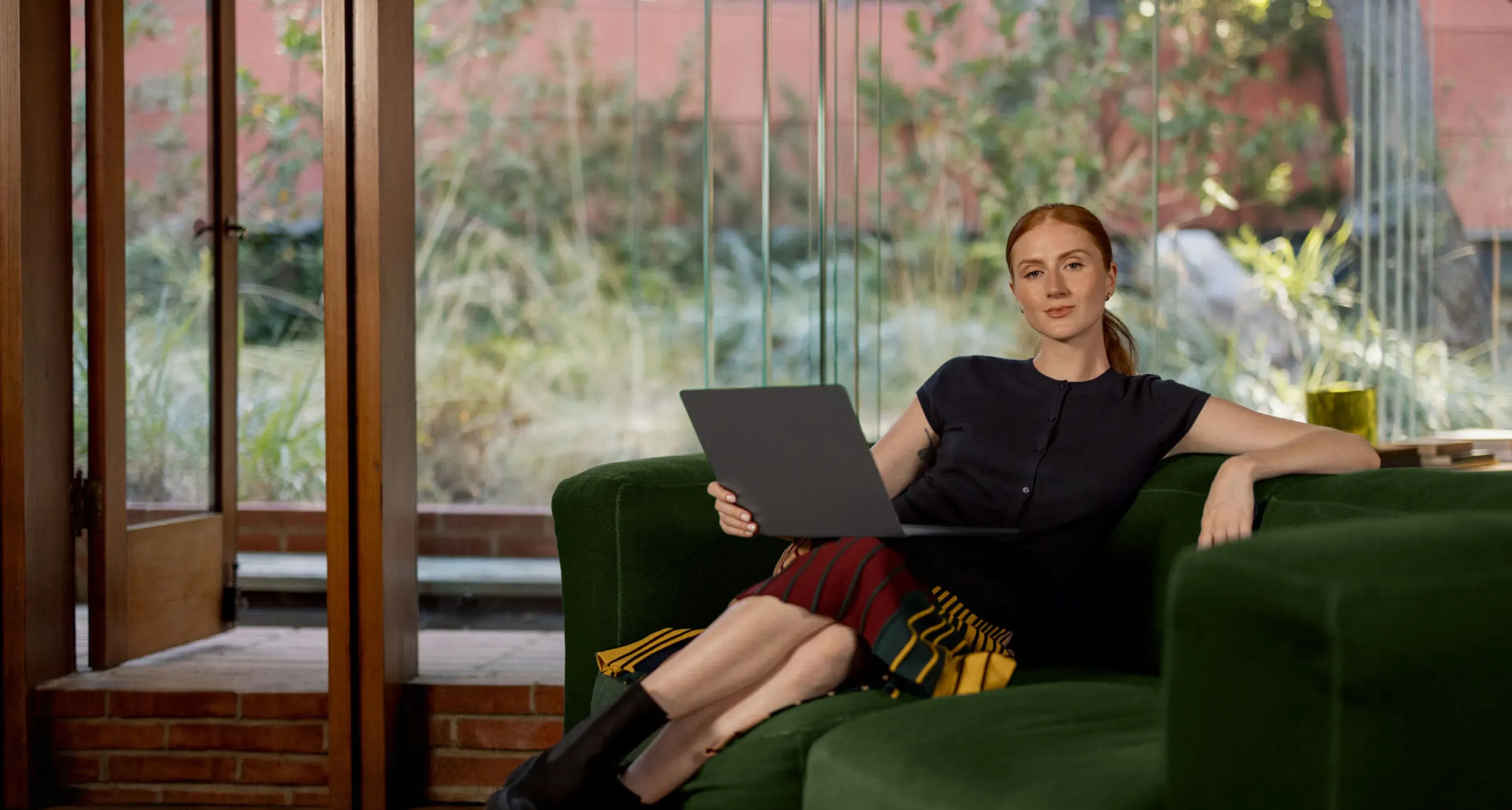 A woman with red hair sits on a green couch, holding a laptop, looking at the camera with large windows and greenery behind her.