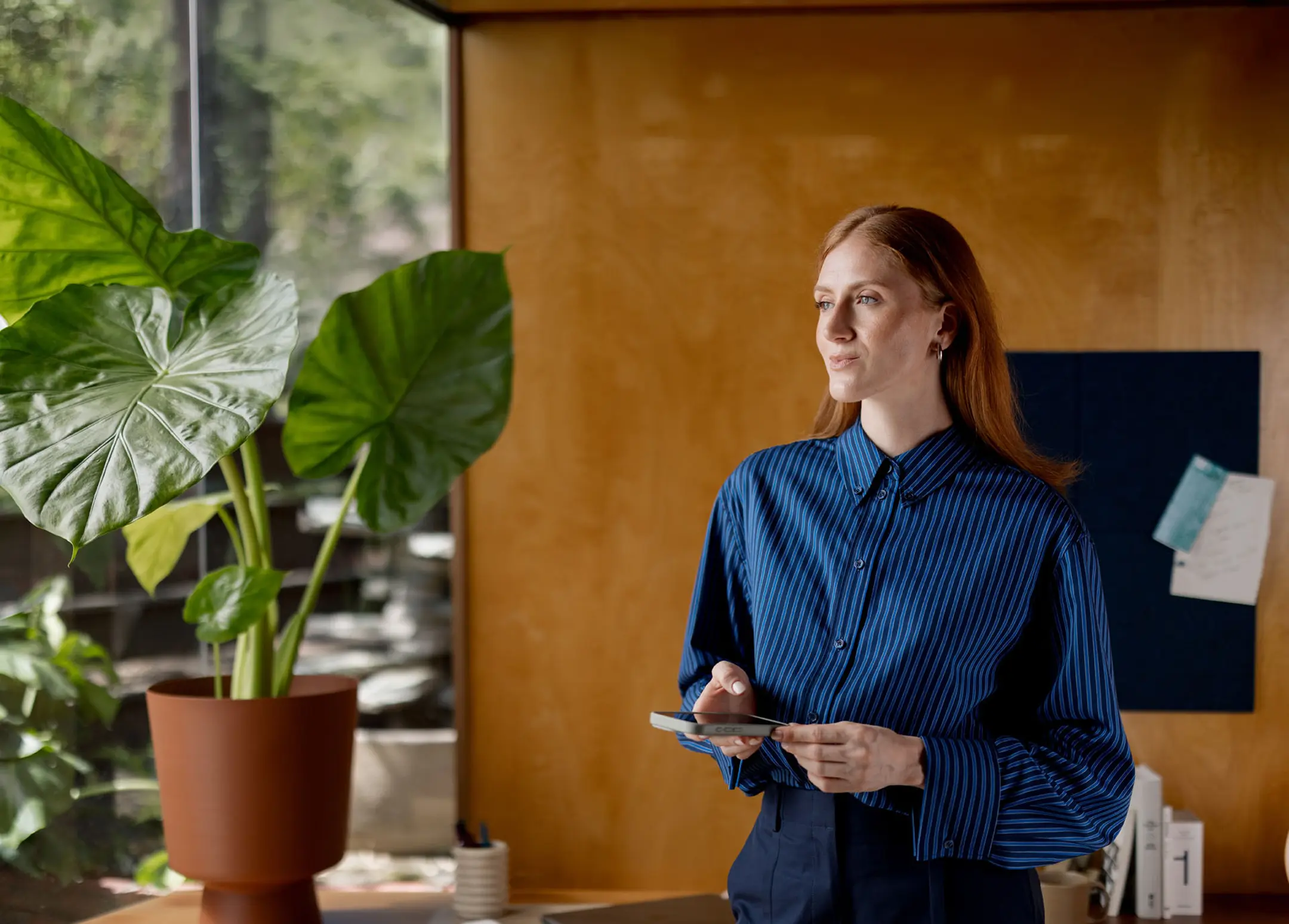Redhead in blue striped shirt holding phone, looking thoughtfully past large potted plant in an indoor office.