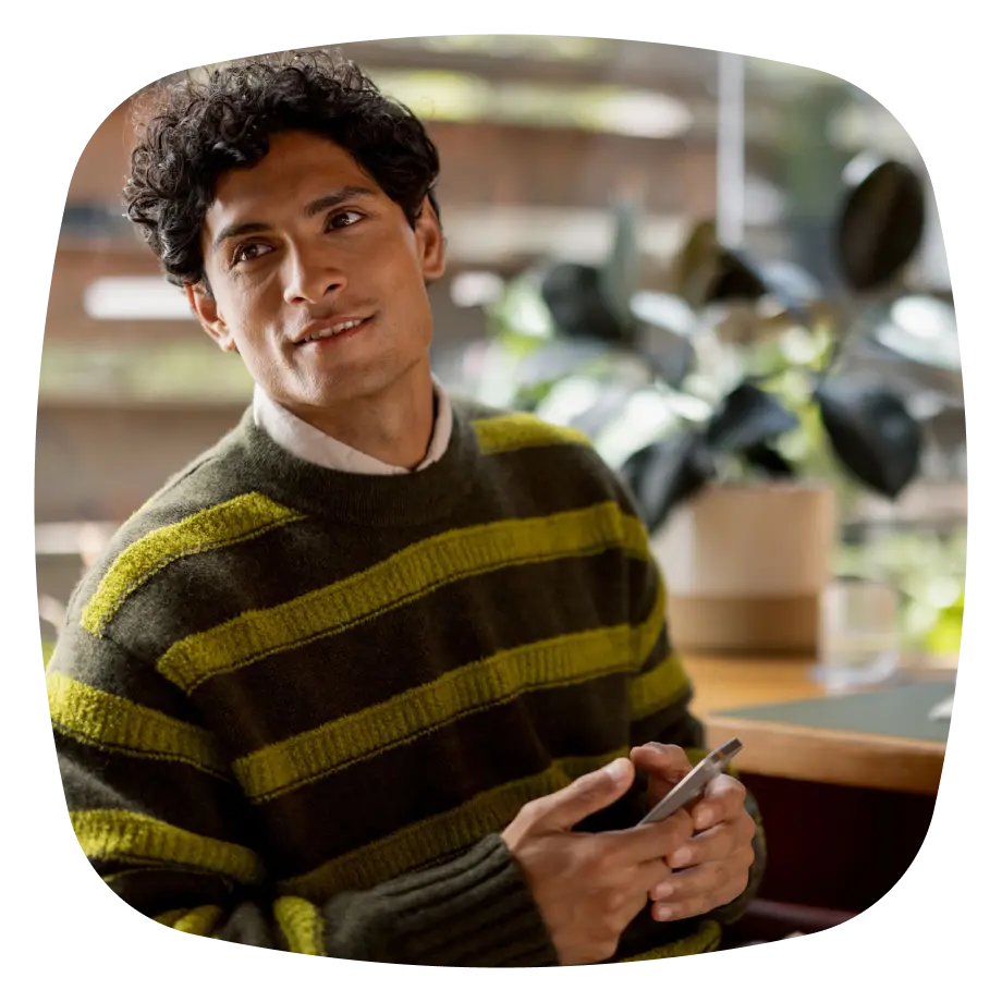 A man with curly dark hair wearing a green and olive striped sweater smiles while holding a smartphone.