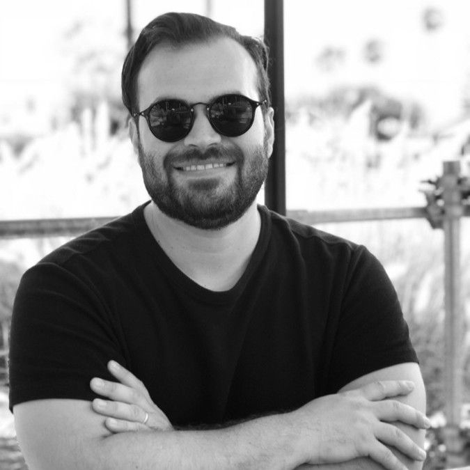 A smiling man with a beard and round sunglasses wears a black t-shirt, arms crossed, in a black and white photo.