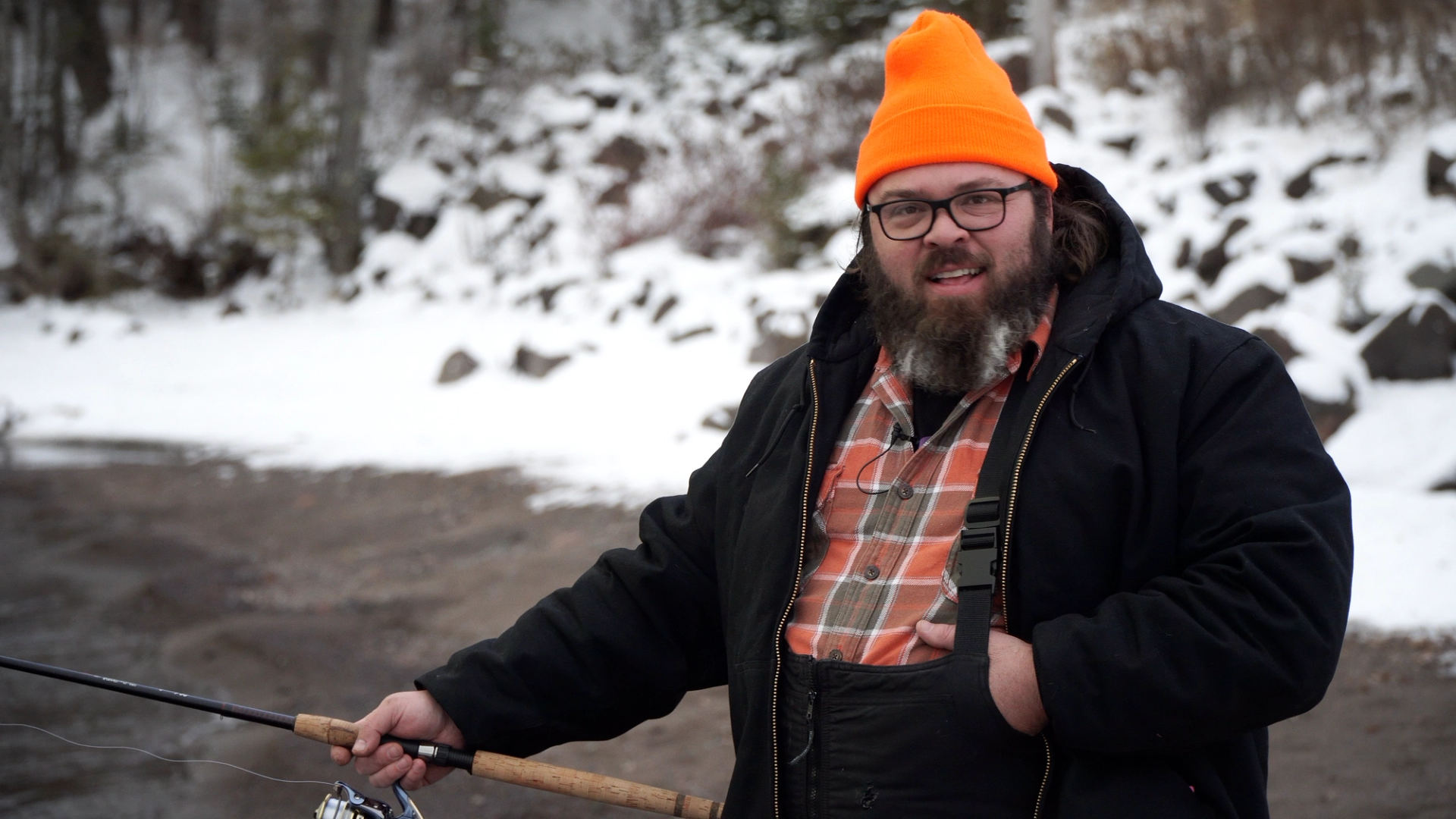 Nate P, in an orange beanie and holding a fishing pole, in front of a snowy river