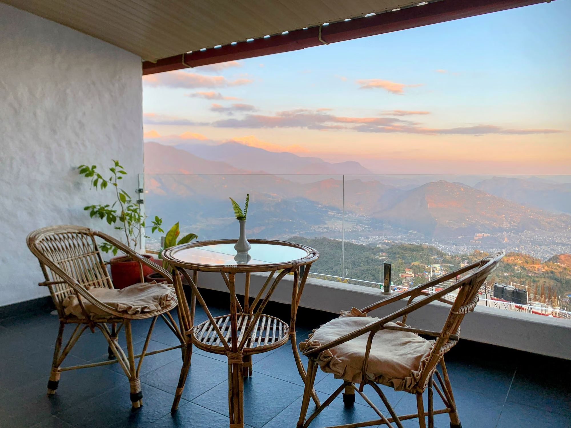 Two cane chairs and a glass table on a balcony over the Pokhara valley at sunset