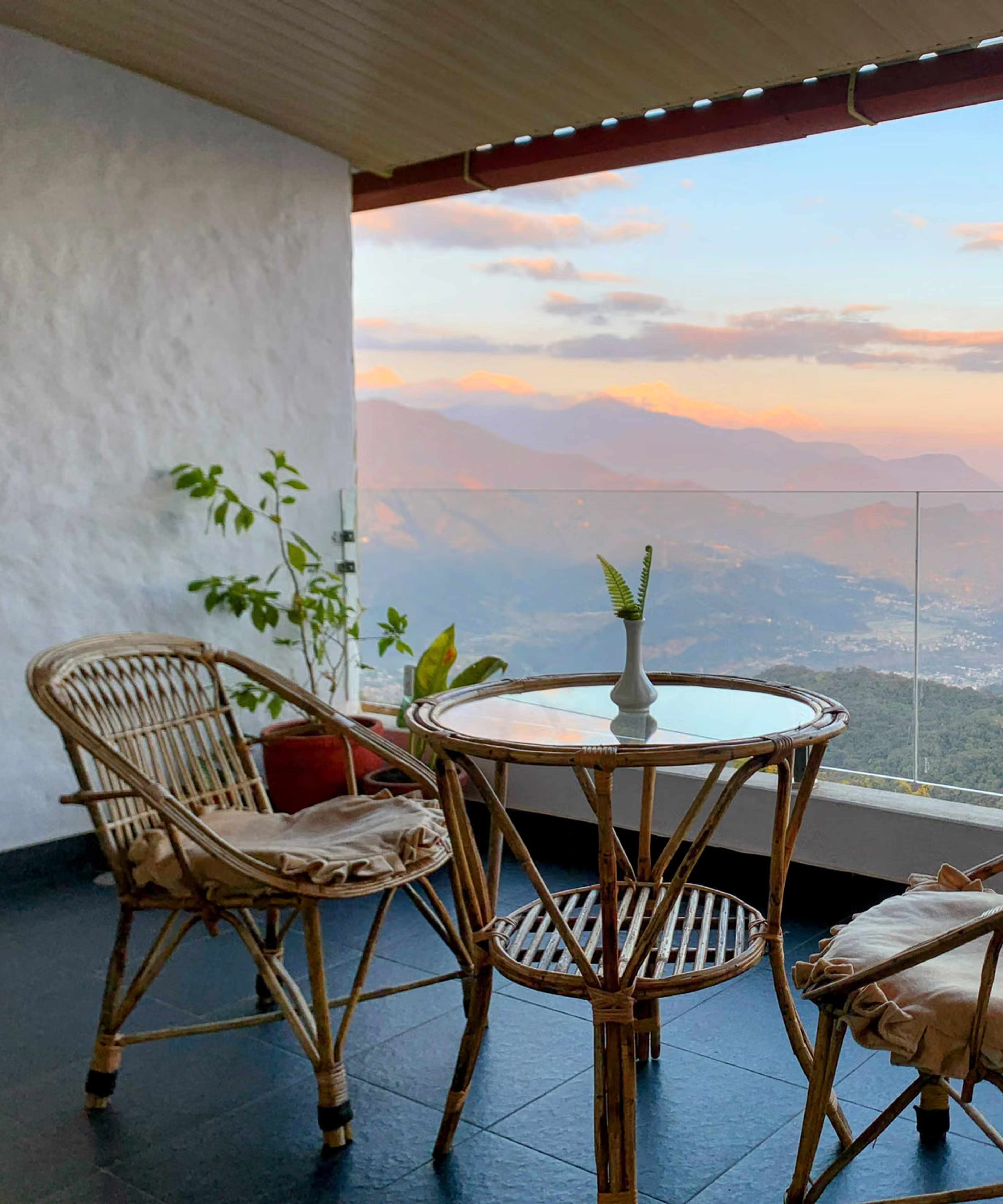 Two cane chairs and a glass table on a balcony over the Pokhara valley at sunset