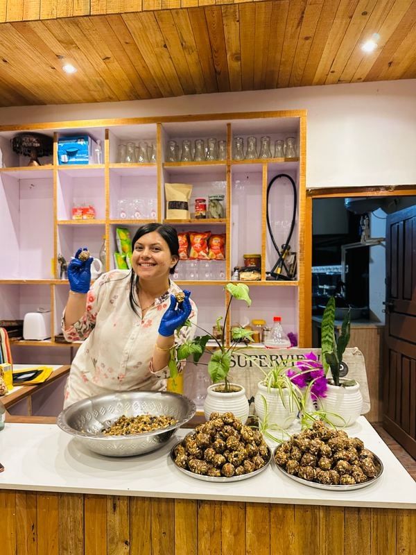 Suraksha in gloves, smiling and holding up two dates and nuts bites, with trays of them on the counter