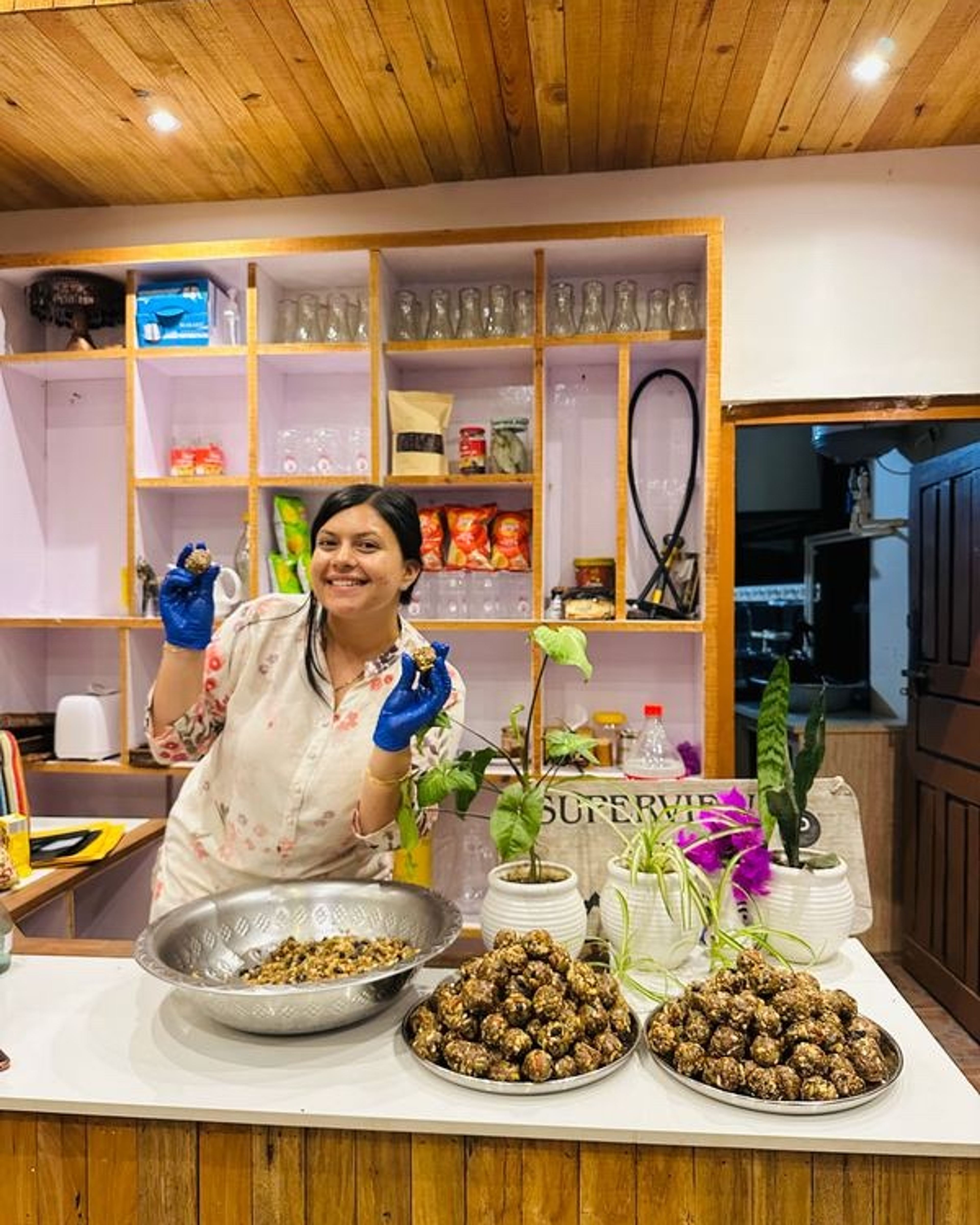 Suraksha in gloves, smiling and holding up two dates and nuts bites, with trays of them on the counter