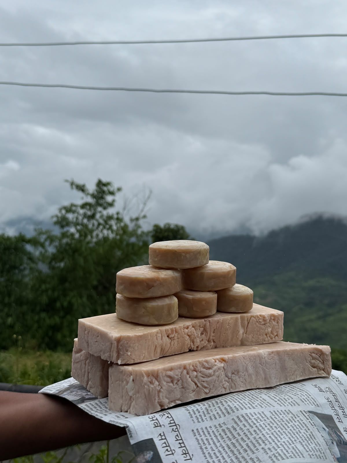Handmade soaps stacked on a newspaper, with cloudy green hills behind