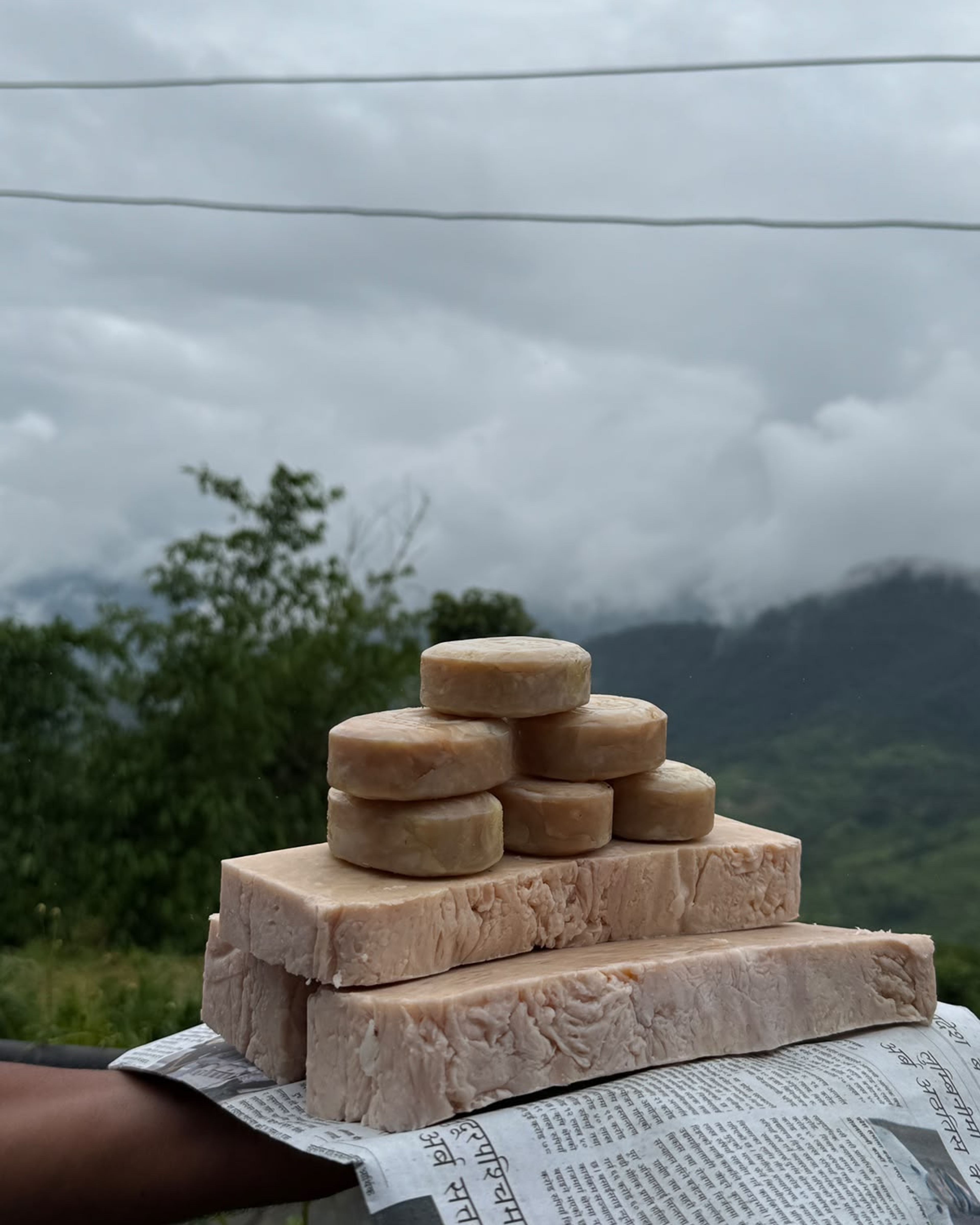 Handmade soaps stacked on a newspaper, with cloudy green hills behind