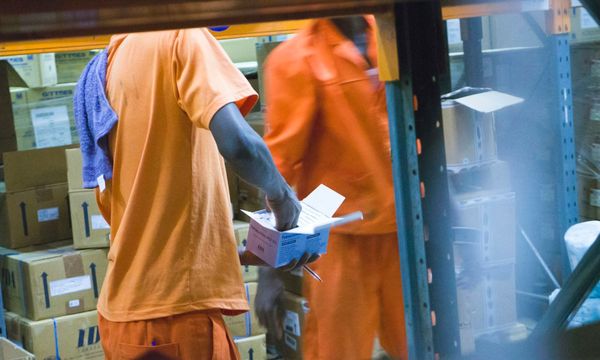 Dua pekerja gudang di depan rak kardus, satu memegang kotak terbuka sambil mencatat jumlah barang di secarik kertas, menggambarkan pencatatan stok manual yang terpisah dari sistem kasir