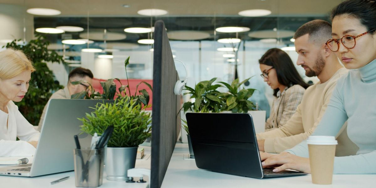 A full open-plan office with a dozen people working on laptops at shared desks, the peak-hour load that makes office Wi-Fi slow