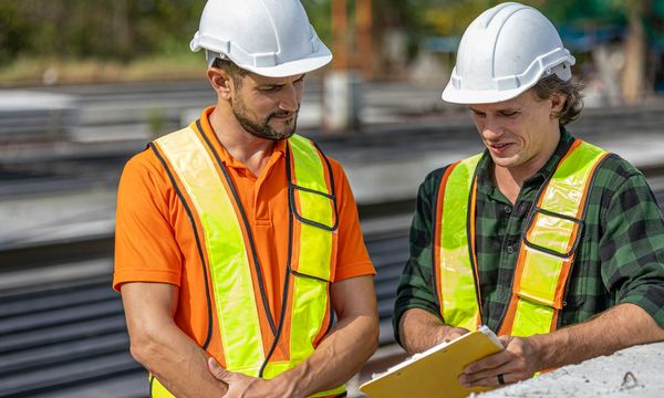Two inspectors in safety vests and hard hats reviewing a checklist on a clipboard, standing in for a security audit team at work