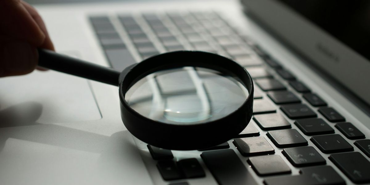 Tangan memegang kaca pembesar di atas papan ketik laptop, menggambarkan pemeriksaan portofolio vendor website satu per satu sebelum memilih