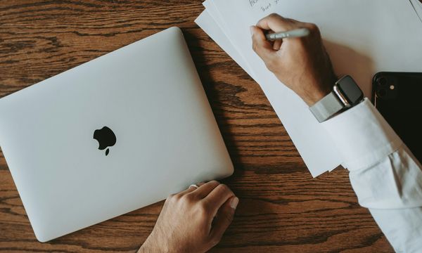 Tampak atas meja kerja dengan tumpukan dokumen kertas yang sedang ditulisi pulpen, laptop tertutup di sebelah kiri dan ponsel di sebelah kanan, menggambarkan dokumen yang akan dibubuhi e-Meterai secara elektronik