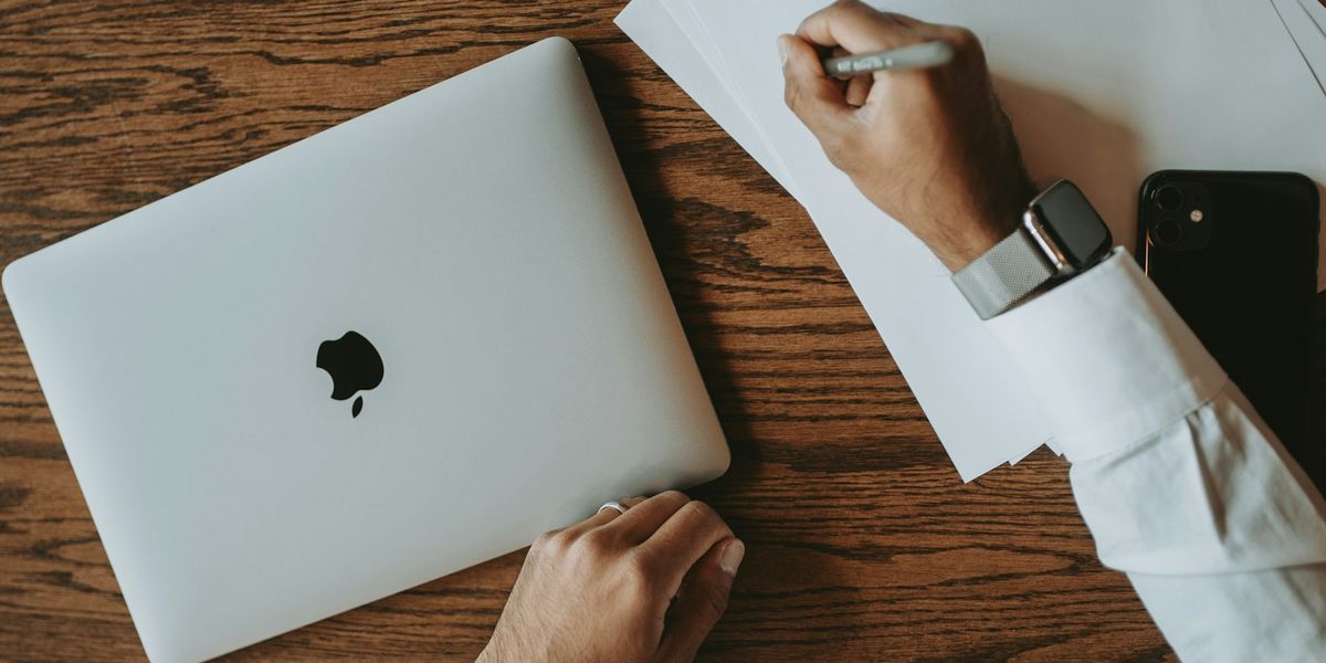 Tampak atas meja kerja dengan tumpukan dokumen kertas yang sedang ditulisi pulpen, laptop tertutup di sebelah kiri dan ponsel di sebelah kanan, menggambarkan dokumen yang akan dibubuhi e-Meterai secara elektronik