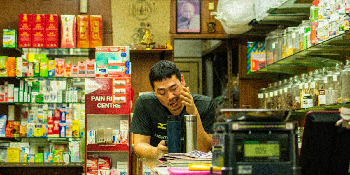 Pemilik toko kelontong duduk di balik meja kasir di antara rak dan toples barang dagangan, dengan mesin kasir lama di depannya