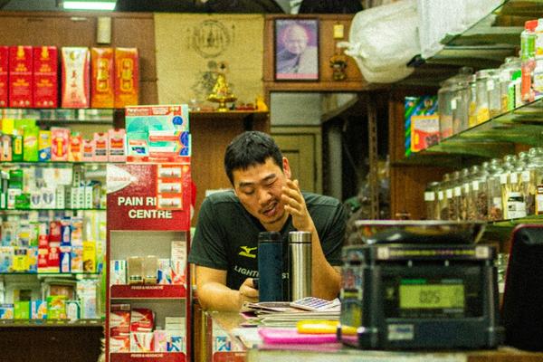 Pemilik toko kelontong duduk di balik meja kasir di antara rak dan toples barang dagangan, dengan mesin kasir lama di depannya