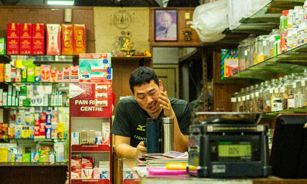 Pemilik toko kelontong duduk di balik meja kasir di antara rak dan toples barang dagangan, dengan mesin kasir lama di depannya